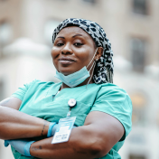 A healthcare professional in scrubs and a patterned headscarf stands outdoors with arms crossed, wearing gloves and a face mask around their neck, ID badge visible.